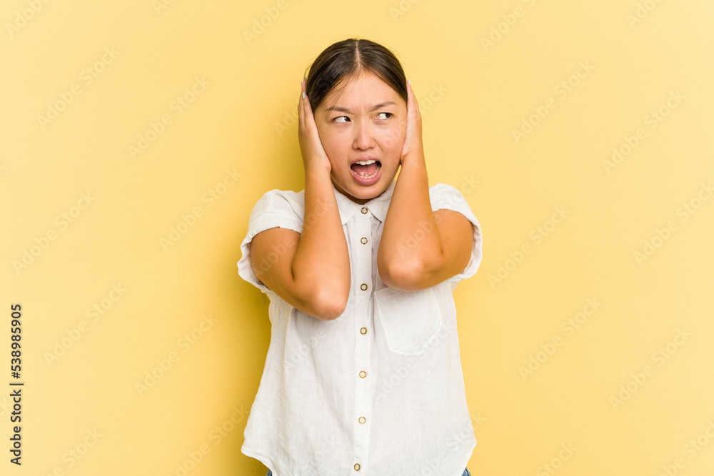 Young Asian woman isolated on yellow background covering ears with hands trying not to hear too loud sound. Young Asian woman isolated on yellow background covering ears with hands trying not to hear too loud sound.