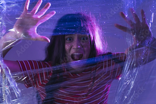 Close up face of a young, caucasian, scared girl with long dark hair looking at the camera through plastic wrap, shouting and having her arms up. Purple led lights. High quality photo
