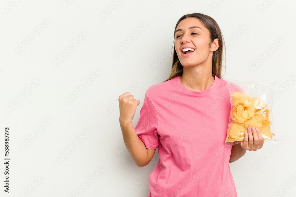 Young Indian woman holding crips isolated on white background raising fist after a victory, winner concept. Young Indian woman holding crips isolated on white background raising fist after a victory, winner concept.