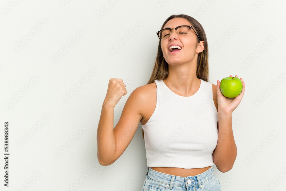 Young Indian woman holding an apple isolated on white background raising fist after a victory, winner concept. Young Indian woman holding an apple isolated on white background raising fist after a victory, winner concept.