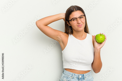 Young Indian woman holding an apple isolated on white background touching back of head, thinking and making a choice.