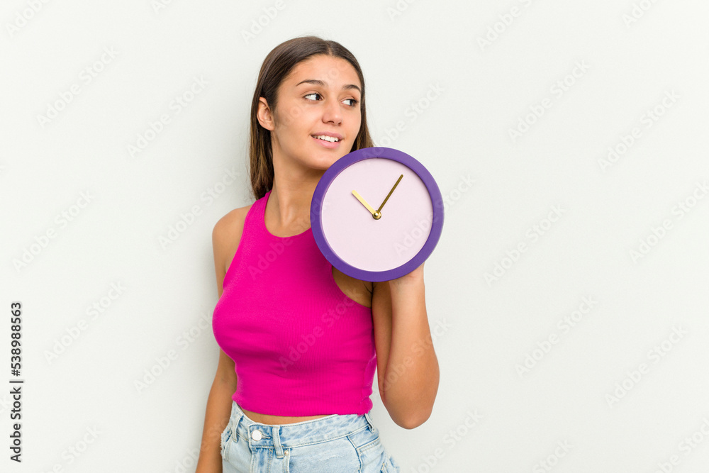 Young Indian woman holding a clock isolated on white background looks aside smiling, cheerful and pleasant. Young Indian woman holding a clock isolated on white background looks aside smiling, cheerful and pleasant.