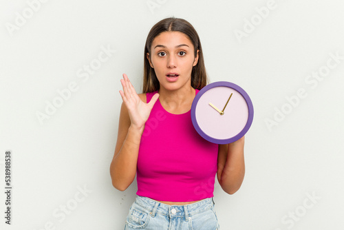 Young Indian woman holding a clock isolated on white background surprised and shocked. © Asier