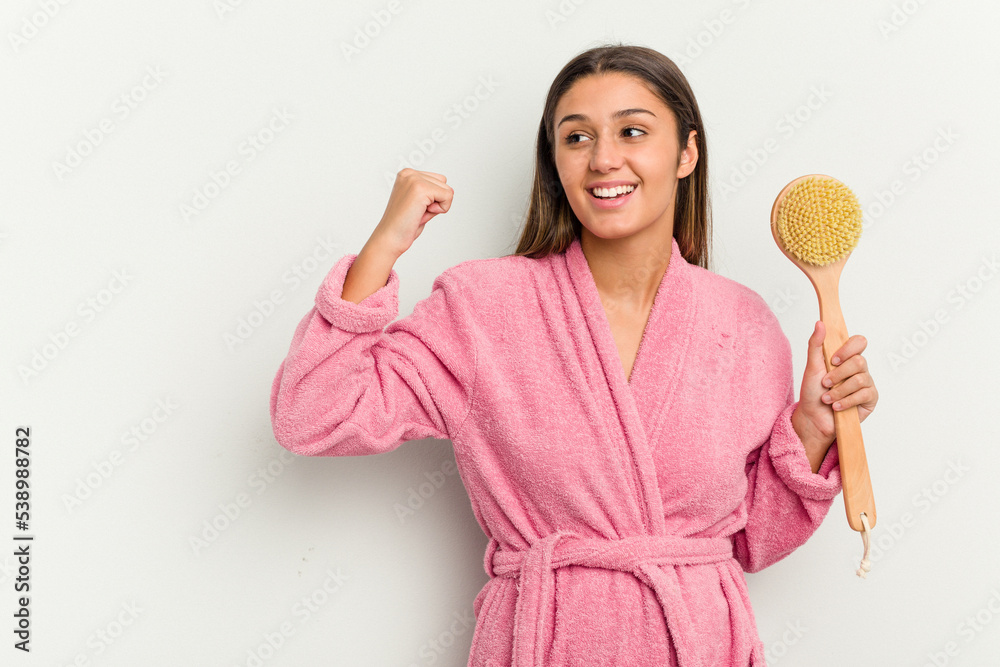 Young Indian woman holding a shower brush isolated on white background raising fist after a victory, winner concept. Young Indian woman holding a shower brush isolated on white background raising fist after a victory, winner concept.