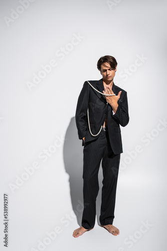 full length of barefoot and brunette man in trendy suit holding pearls while posing with hand in pocket on grey