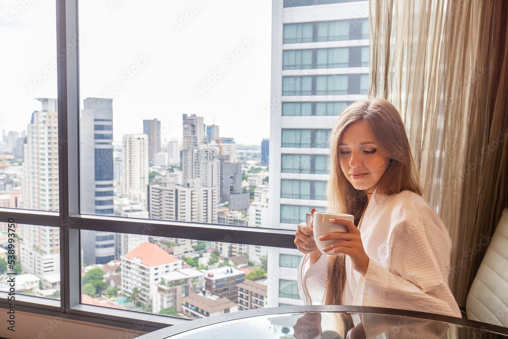 Fototapeta premium Woman in bathrobe with coffee cup sitting in modern hotel or apartment