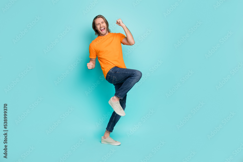 Full length photo of charming lucky guy dressed orange t-shirt smiling rising fists isolated teal color background