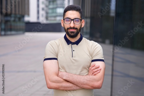Portrait of happy positive confident successful Arab African Muslim ethnic guy, young bearded businessman, man with black beard standing outdoors smiling looking at camera with his hands crossed