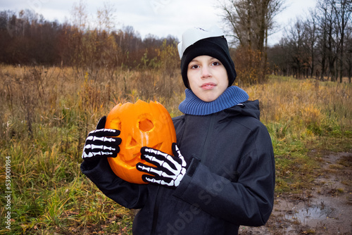 Child in skeleton gloves holding halloween pumpkin jack-o'-lantern on background of trees