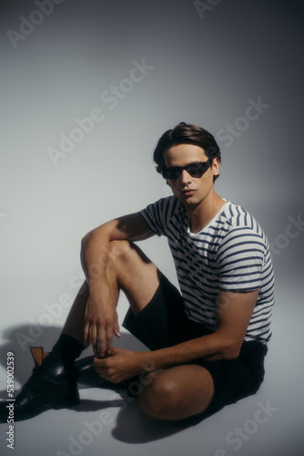 Stylish man in shorts and shoes sitting on grey background with shadow