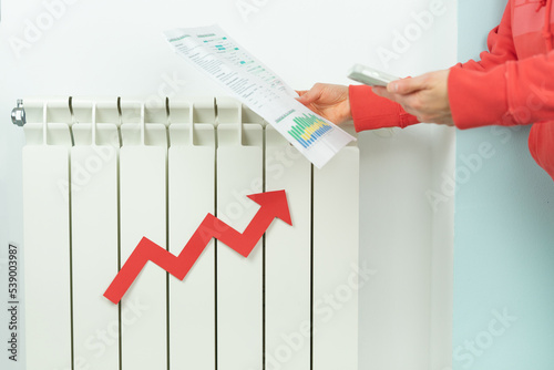 Hands with electricity bill closing heating radiator due to price hike. Energy crisis.