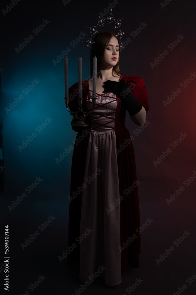 Mythical fantasy queen with a candlestick. Silvery gothic crown on the head. Burgundy vintage artistic dress, black gloves. Medieval style. Full-length portrait in a dark key. Vertical photo. Mythical fantasy queen with a candlestick. Silvery gothic crown on the head. Burgundy vintage artistic dress, black gloves. Medieval style. Full-length portrait in a dark key. Vertical photo.