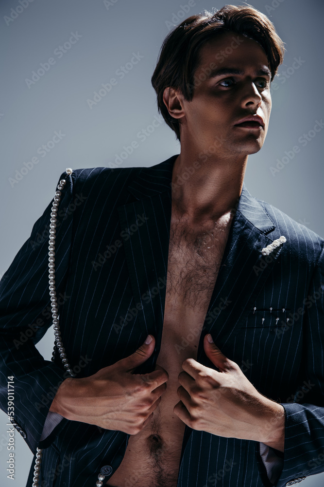 brunette man with hairy chest adjusting stylish blazer with pearl chain on  dark grey Photos | Adobe Stock