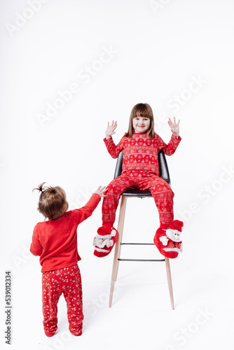 Merry Christmas and Happy family Holidays. Cheerful cute children girls wearing in Xmas red pajames having fun on white background. Smiling older sister sitting on chair and playing. Loving sibling photo