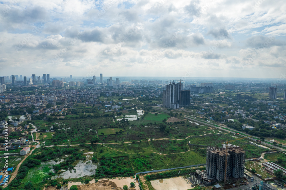 aerial drone shot over gurgaon showing monsoon clouds with light rays falling on ground crowded with homes houses, sohna highway feilds and water pools around under construction buildings