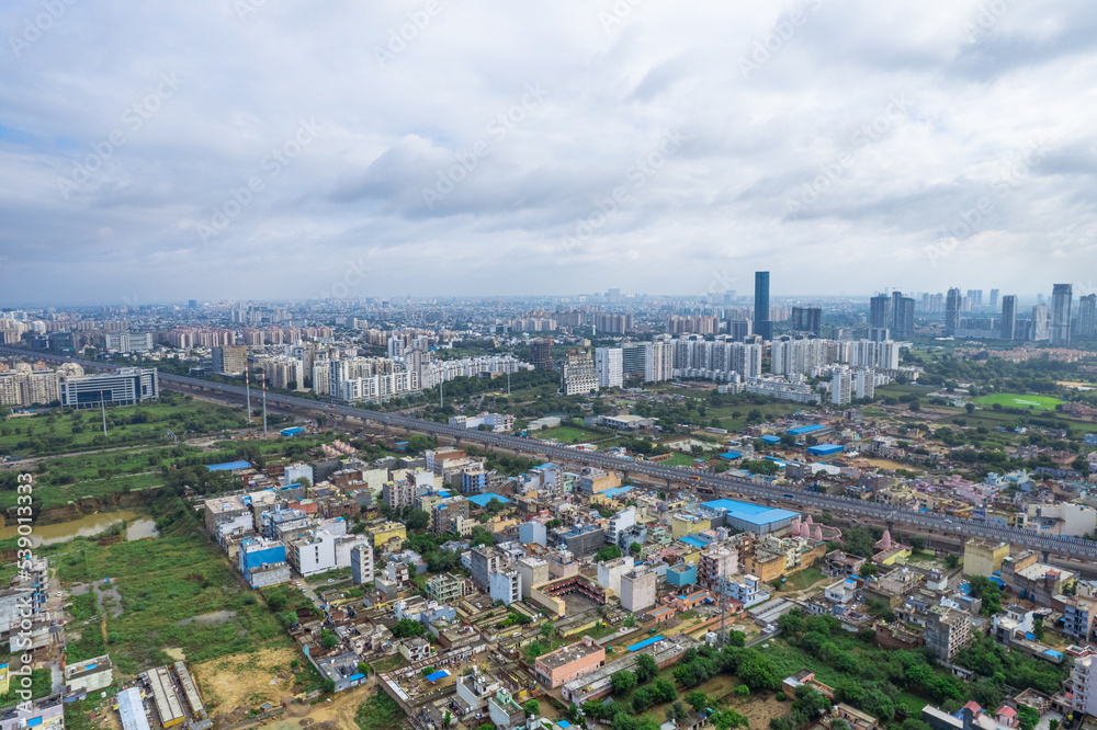 Fototapeta premium aerial drone shot over gurgaon showing monsoon clouds with light rays falling on ground crowded with homes houses, sohna highway feilds and water pools around under construction buildings
