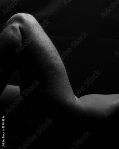 Artistic male nude body fragment close up of raised bare thigh or leg and athletic man torso, sexy figure on black background, vertical