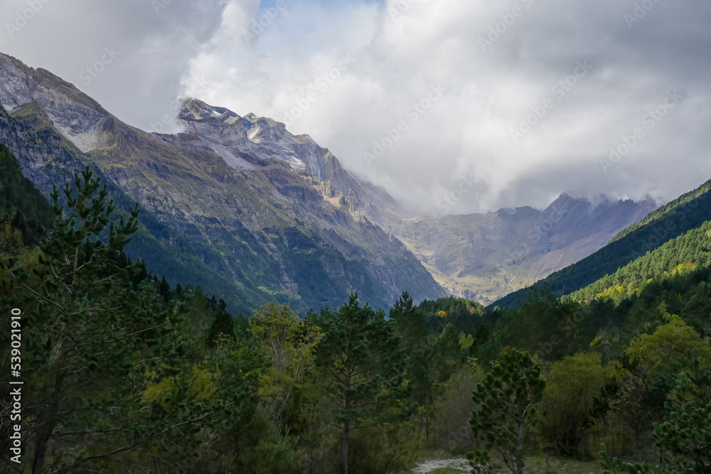 view along a glacial forest lined valley to a massive mountainous amphitheater (cirque) with cloud forming on the mountain tops view along a glacial forest lined valley to a massive mountainous amphitheater (cirque) with cloud forming on the mountain tops