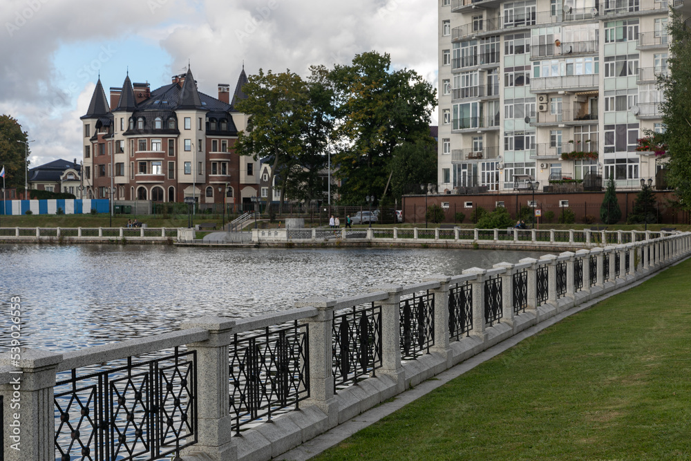 Naklejka premium City embankment of Upper Pond in Kaliningrad in the cloudy day. Curly white stone and metal fence along the river. Rich residential building on the opposite riverbank. People are walking on the quay