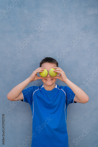 Portrait of a young boy holding tennis balls over his eyes on blue background with copy space. Sports concept © diignat