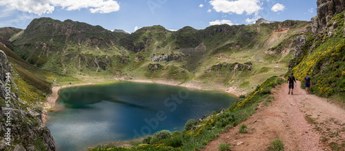 Una persona caminando en un paisaje asturiano, por un sendero del Lago de la Cueva rodeado de naturaleza, montañas verdes, flores moradas cielo azul y nubes blancas en verano de 2021, España.