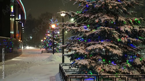 Christmas trees decorated with garlands and balloons on the street in a snowfall, pedestrians are walking on the street