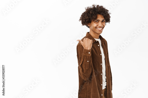 Portrait of handsome young hispanic man pointing finger left, behind his back, showing logo or banner, chart on copy space, standing over white background