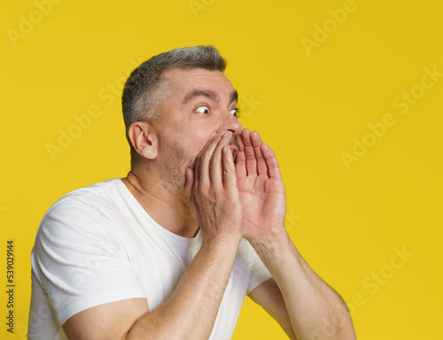 Screaming loud handsome grey haired man cover his mouth with hands to help send sound wearing white t-shirt isolated on yellow background. Spreading a word man whispering or gossiping