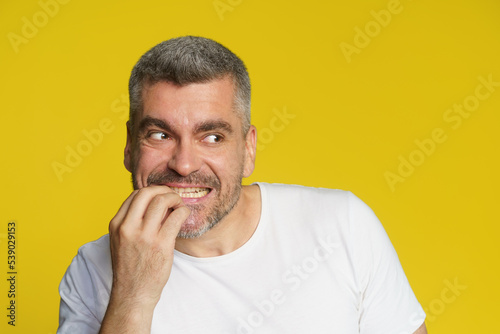 Scared of competitor handsome middle aged man bitting his fingernails. Freelancer in stress of project deadline. Shocked freelancer man eating fingers in white t-shirt isolated on yellow background