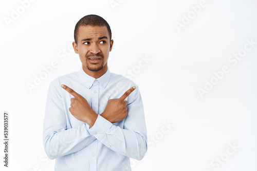 Portrait of confused office worker, young man pointing sideways, cannot make decision, look clueless, standing over white background