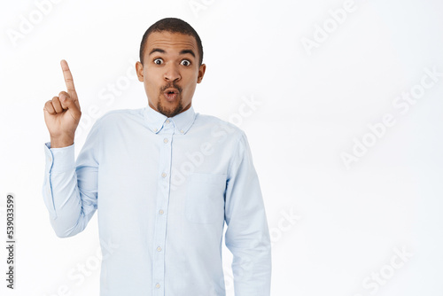 Surprised businessman pointing finger up and say wow with impressed face, standing amazed over white background photo