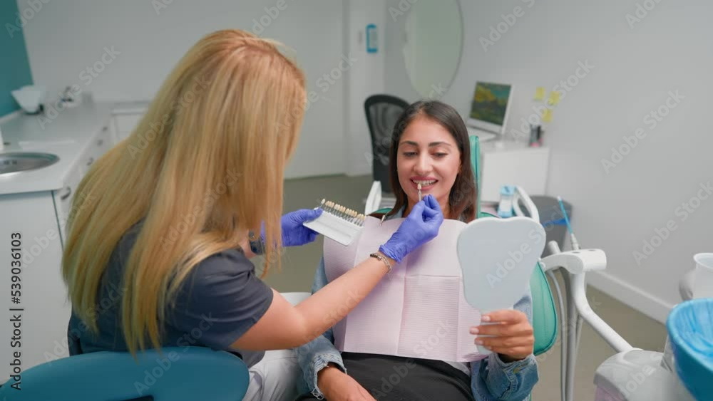Woman Professional Dentist Examines a Smiling Female Patient in a Dental Clinic. Selects the Shade of Dental Implants Before Operation. Concept of Healthcare and Medicine. Slow Motion. Woman Professional Dentist Examines a Smiling Female Patient in a Dental Clinic. Selects the Shade of Dental Implants Before Operation. Concept of Healthcare and Medicine. Slow Motion.