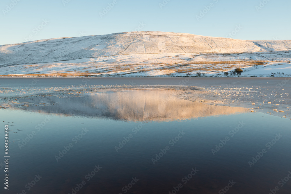 Hills covered in snow reflected on the calm water of one of the reservoir in the Pentland Hills Regional Park in Edinburgh, Scotland, UK, in a cold Winter day with blue skies