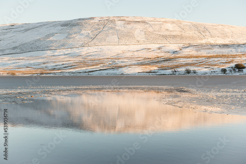 A hill covered in snow reflected on the calm water of one of the reservoir in the Pentland Hills Regional Park in Edinburgh, Scotland, UK, in a cold Winter day with blue skies