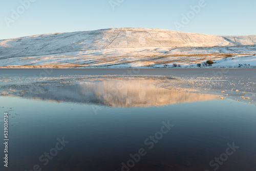 Hills covered in snow reflected on the calm water of one of the reservoir in the Pentland Hills Regional Park in Edinburgh, Scotland, UK, in a cold Winter day with blue skies