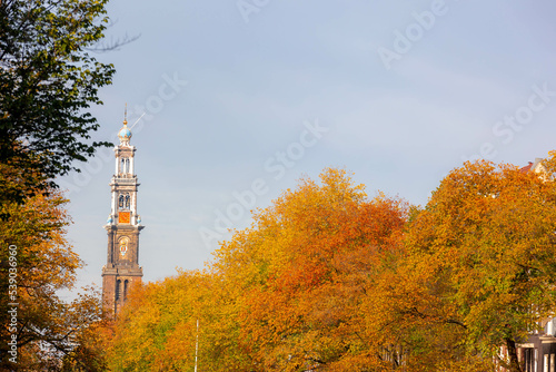 Autumn cityscape in Amsterdam with view of the Westerkerk church tower under blue clear sky, Colourful yellow orange leaves on the trees along Prinsengracht canal, Holland, Netherlands.