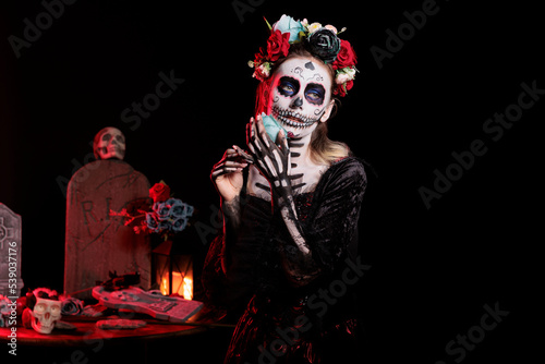 Beautiful woman wearing black and white make up and flowers crown to cleebrate mexican halloween ritual. Goddess of death with horror carnival costume and body art, holding rose.
