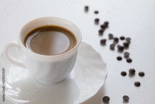 Tazza bianca con caff   e piattino con chicchi di cioccolato su tavolo bianco