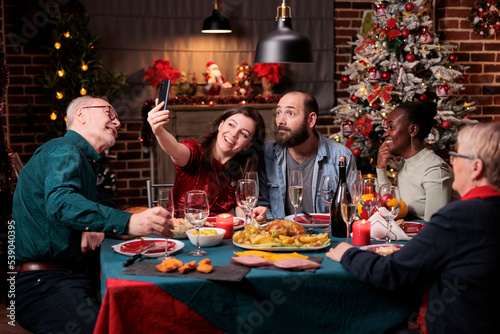 Woman taking smartphone selfie of family christmas celebration  friends portrait at festive dinner table. Diverse people gathering  celebrating xmas  eating traditional meal