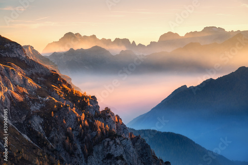 Mountains in low clouds at beautiful sunset in autumn in Dolomites, Italy. Landscape with alpine mountain hills in fog, orange trees and grass in fall, colorful sky with golden sunbeams. Aerial view © den-belitsky