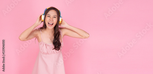 Happy asian woman wear headphones listening playlist musing standing on pink background. Excited young girl listen sound melody radio with earphones song music over isolated pastel background. Hobby