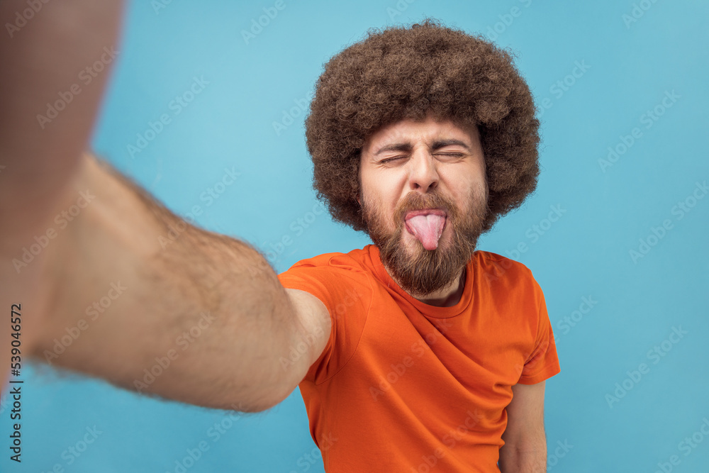 Portrait of childish silly man with Afro hairstyle wearing orange T-shirt taking selfie or has livestream, showing tongue out, point of view photo. Indoor studio shot isolated on blue background. Portrait of childish silly man with Afro hairstyle wearing orange T-shirt taking selfie or has livestream, showing tongue out, point of view photo. Indoor studio shot isolated on blue background.