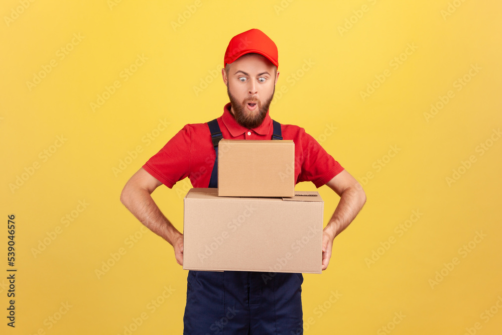 Amazed surprised deliveryman in uniform holding two heavy big cardboard boxes in hands, having funny facial expression, delivering oreder. Indoor studio shot isolated on yellow background. Amazed surprised deliveryman in uniform holding two heavy big cardboard boxes in hands, having funny facial expression, delivering oreder. Indoor studio shot isolated on yellow background.