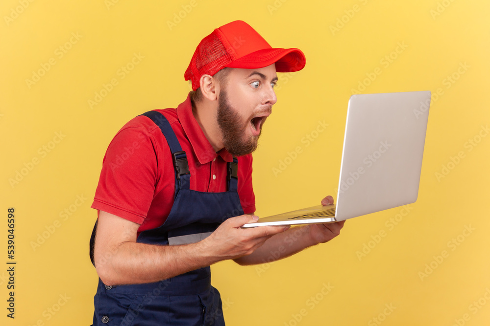 Side view of extremely shocked handyman standing laptop in hands and looking at notebook display with big eyes, wearing uniform and red cap. Indoor studio shot isolated on yellow background.