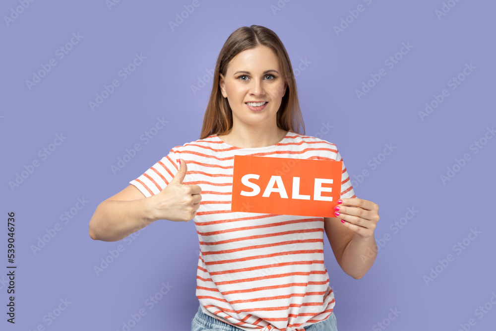 Portrait of beautiful satisfied woman wearing striped T-shirt standing with sale card in hands and showing thumb up, like gesture, like good discounts. Indoor studio shot isolated on purple background Portrait of beautiful satisfied woman wearing striped T-shirt standing with sale card in hands and showing thumb up, like gesture, like good discounts. Indoor studio shot isolated on purple background