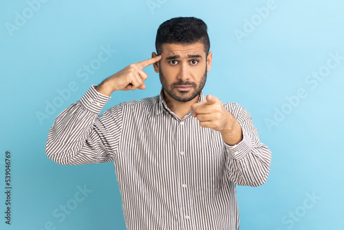 Businessman twirling finger to temple pointing finger at camera, saying you are dumb and stupid, dissatisfied with dumbness, wearing striped shirt. Indoor studio shot isolated on blue background. photo
