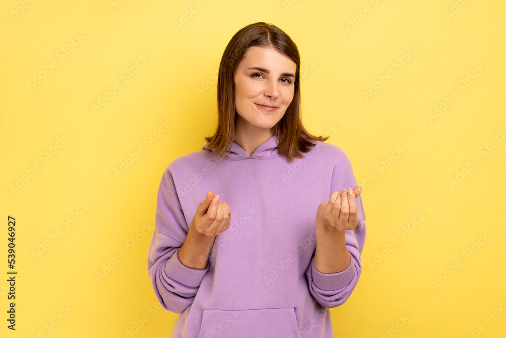 Attractive smiling young adult woman standing and looking at camera with money or italian gesture with hand, wearing purple hoodie. Indoor studio shot isolated on yellow background. Attractive smiling young adult woman standing and looking at camera with money or italian gesture with hand, wearing purple hoodie. Indoor studio shot isolated on yellow background.