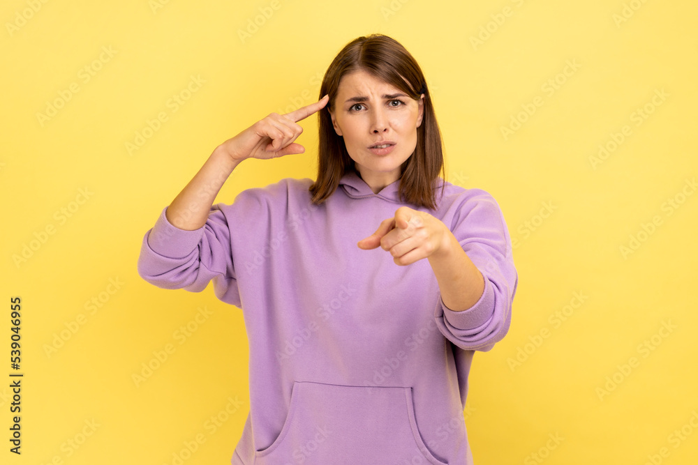 You are crazy. Attractive woman with hair holding finger near temple and pointing on you, showing stupid gesture, wearing purple hoodie. Indoor studio shot isolated on yellow background. You are crazy. Attractive woman with hair holding finger near temple and pointing on you, showing stupid gesture, wearing purple hoodie. Indoor studio shot isolated on yellow background.