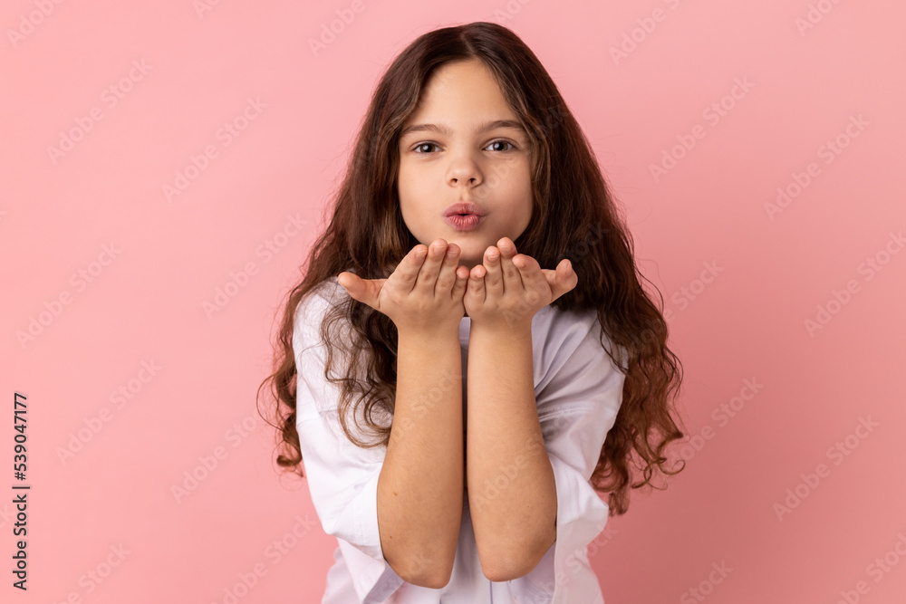 Portrait of romantic lovely little girl wearing white T-shirt sending air kisses to her friend, looking at camera, falling in love. Indoor studio shot isolated on pink background. Portrait of romantic lovely little girl wearing white T-shirt sending air kisses to her friend, looking at camera, falling in love. Indoor studio shot isolated on pink background.