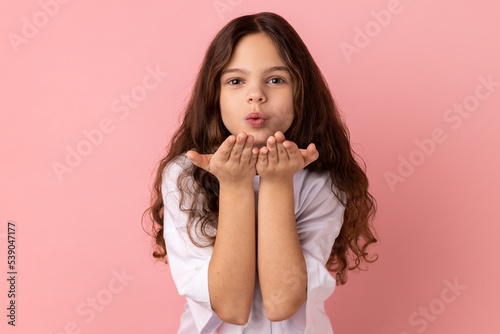 Portrait of romantic lovely little girl wearing white T-shirt sending air kisses to her friend, looking at camera, falling in love. Indoor studio shot isolated on pink background.